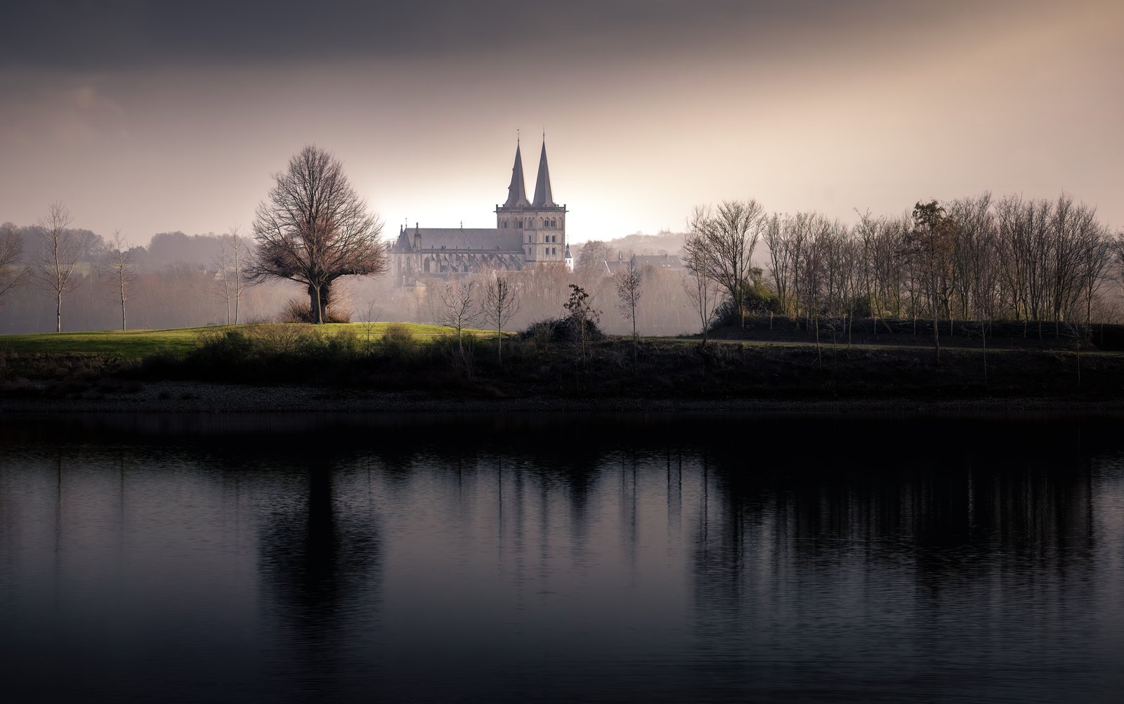 Kirchtürme und winterliche Bäume stehen im Dunst hinter einem dunklen Fluss.