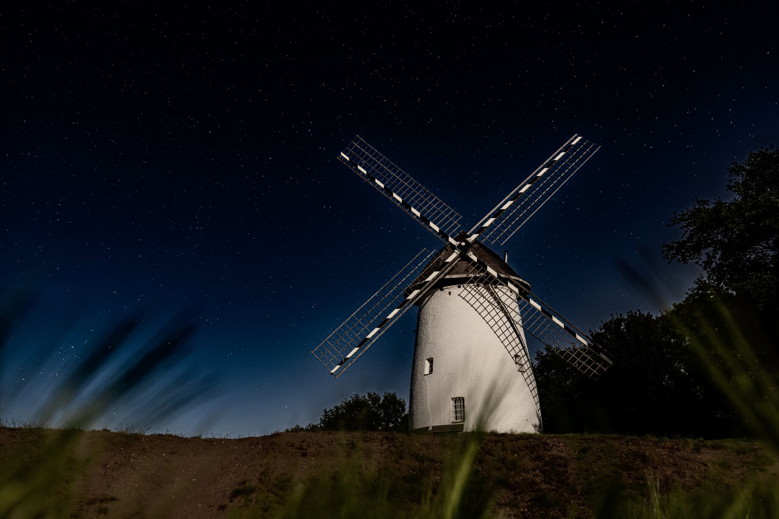 Eine weiße Windmühle steht unter einem tiefblauen Sternenhimmel, langes Gras verwischt im Vordergrund.