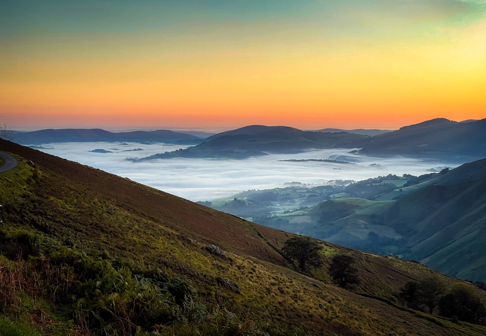 Bergketten und ein Tal mit tiefem Nebel liegen unter einem pastellfarbenen Morgenhimmel.