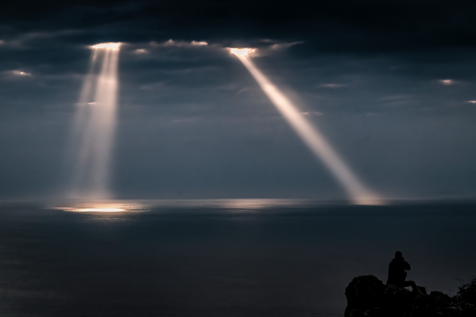 Dunkle Meeresfläche unter schweren Wolken, einzelne Lichtstrahlen auf dem Wasser und eine kleine Person auf dem Fels im Vordergrund.