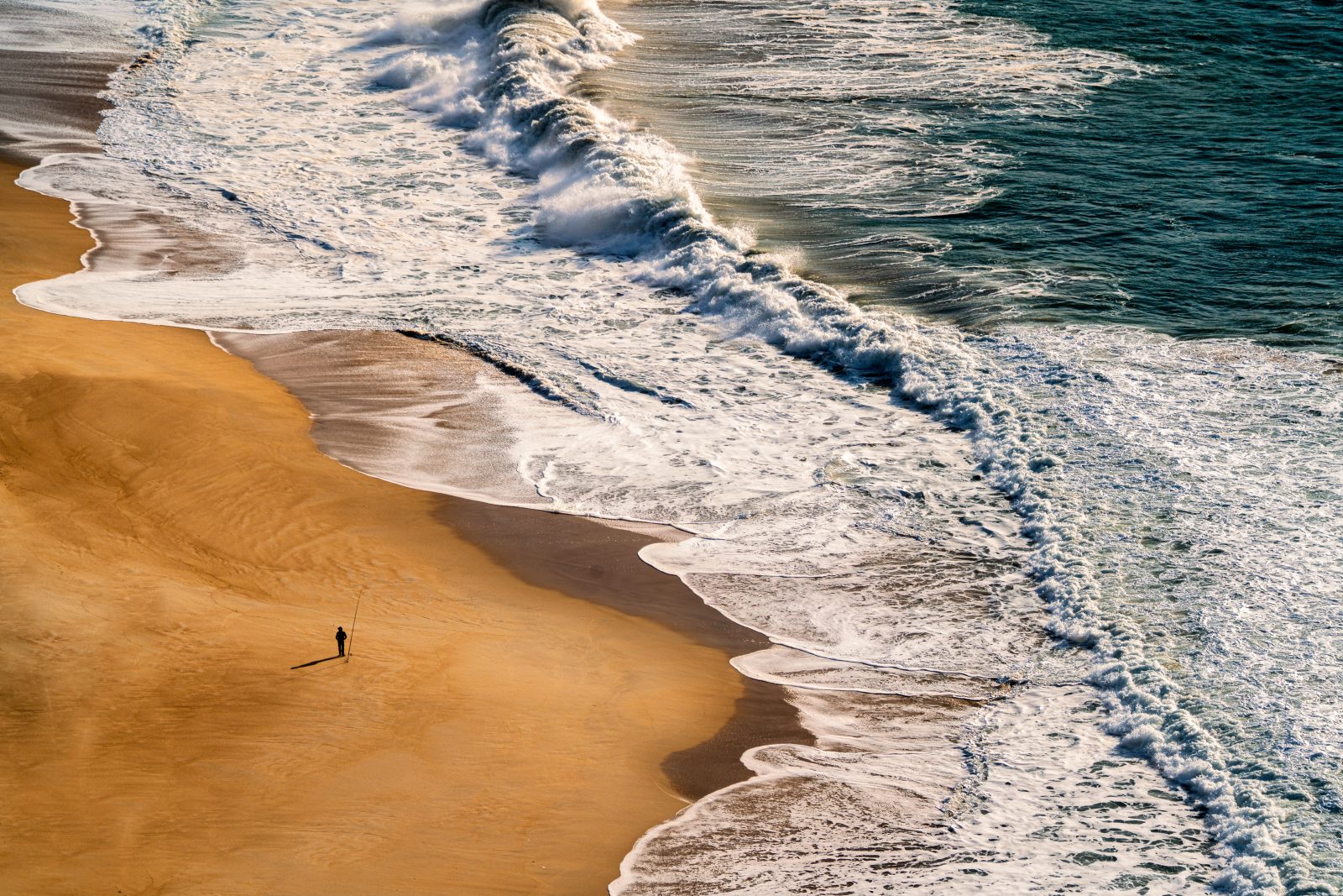 Von oben gesehene Brandung, die über einen breiten Sandstrand läuft; eine einzelne entfernte Person steht nahe der Wasserlinie.