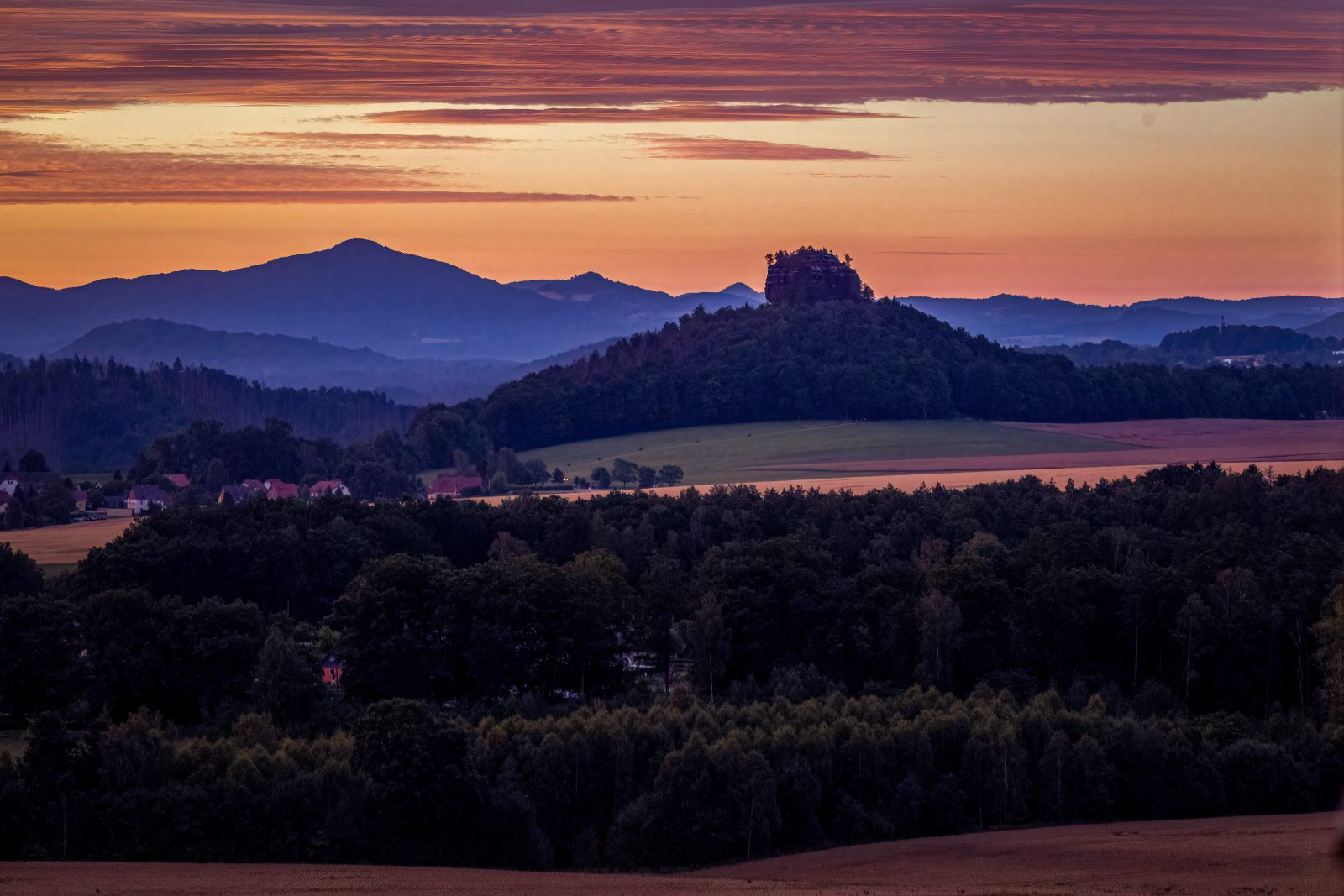 Hügellandschaft mit Feldern, Wald, verstreuten Häusern und einem orangefarbenen Abendhimmel.