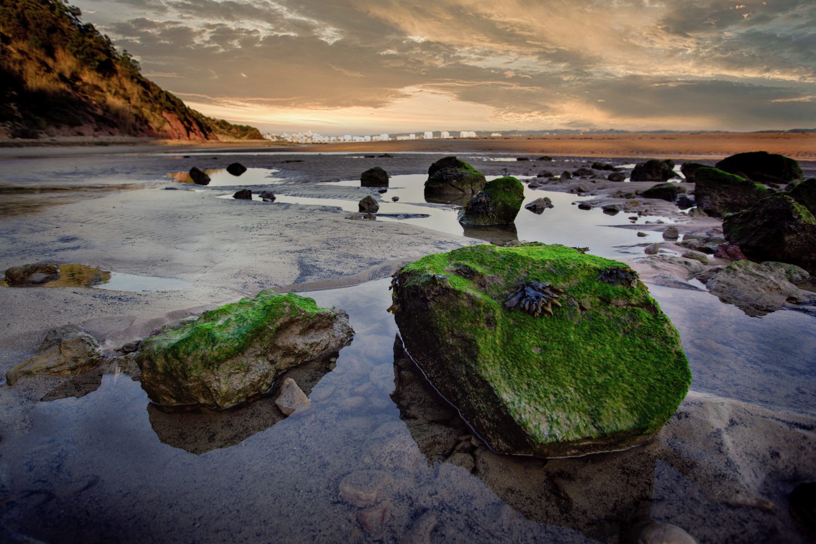 Felsblöcke mit grünem Bewuchs liegen im flachen Wasser eines Gezeitenufers. Im Hintergrund öffnen sich Strand, Felsküste und ein wolkenreicher Himmel im Abendlicht.
