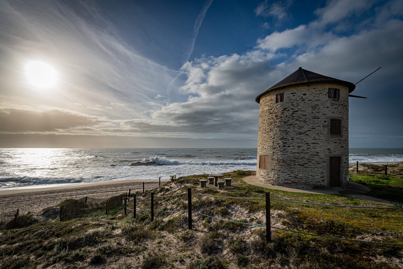 Eine steinerne Windmühle steht auf einer Düne über einem breiten Sandstrand und der bewegten See. Tief stehende Sonne und aufgelockerte Wolken prägen den Himmel.