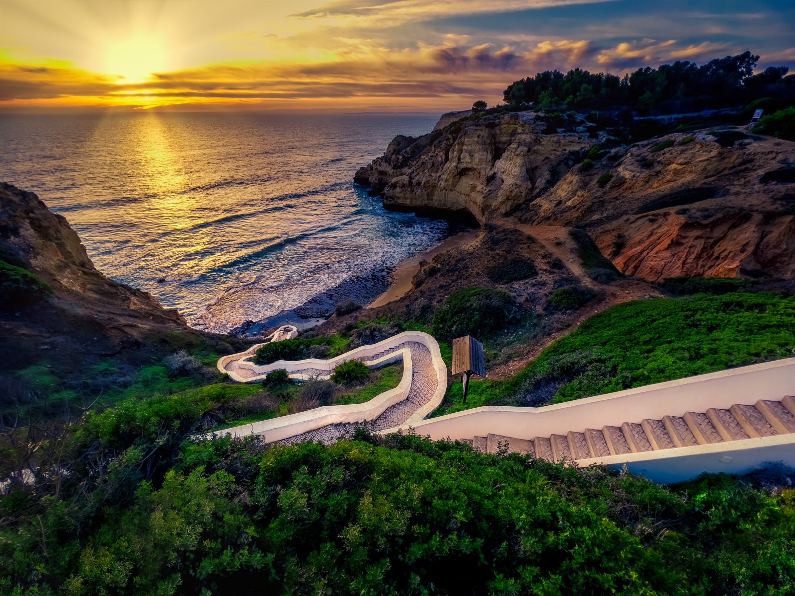 Eine geschwungene Treppe führt über bewachsene Klippen zu einer kleinen Bucht an der Algarve; die tief stehende Sonne spiegelt sich im Atlantik.