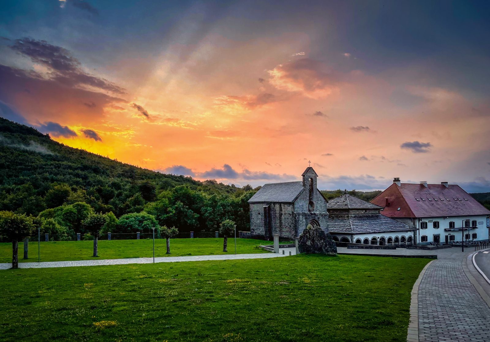 Eine steinerne Kirche und angrenzende Gebäude stehen bei Sonnenuntergang an einem bewaldeten Hang; der Himmel leuchtet orange, rosa und blau.