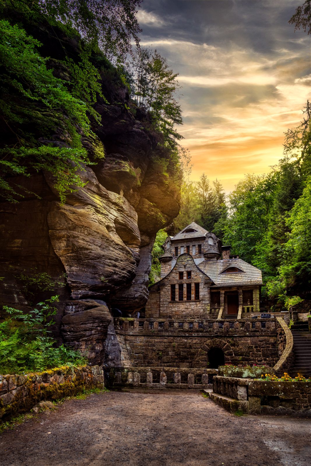 Historisches Steinhaus unter einer markanten Felswand in bewaldeter Umgebung, mit Treppenanlage und Abendhimmel.