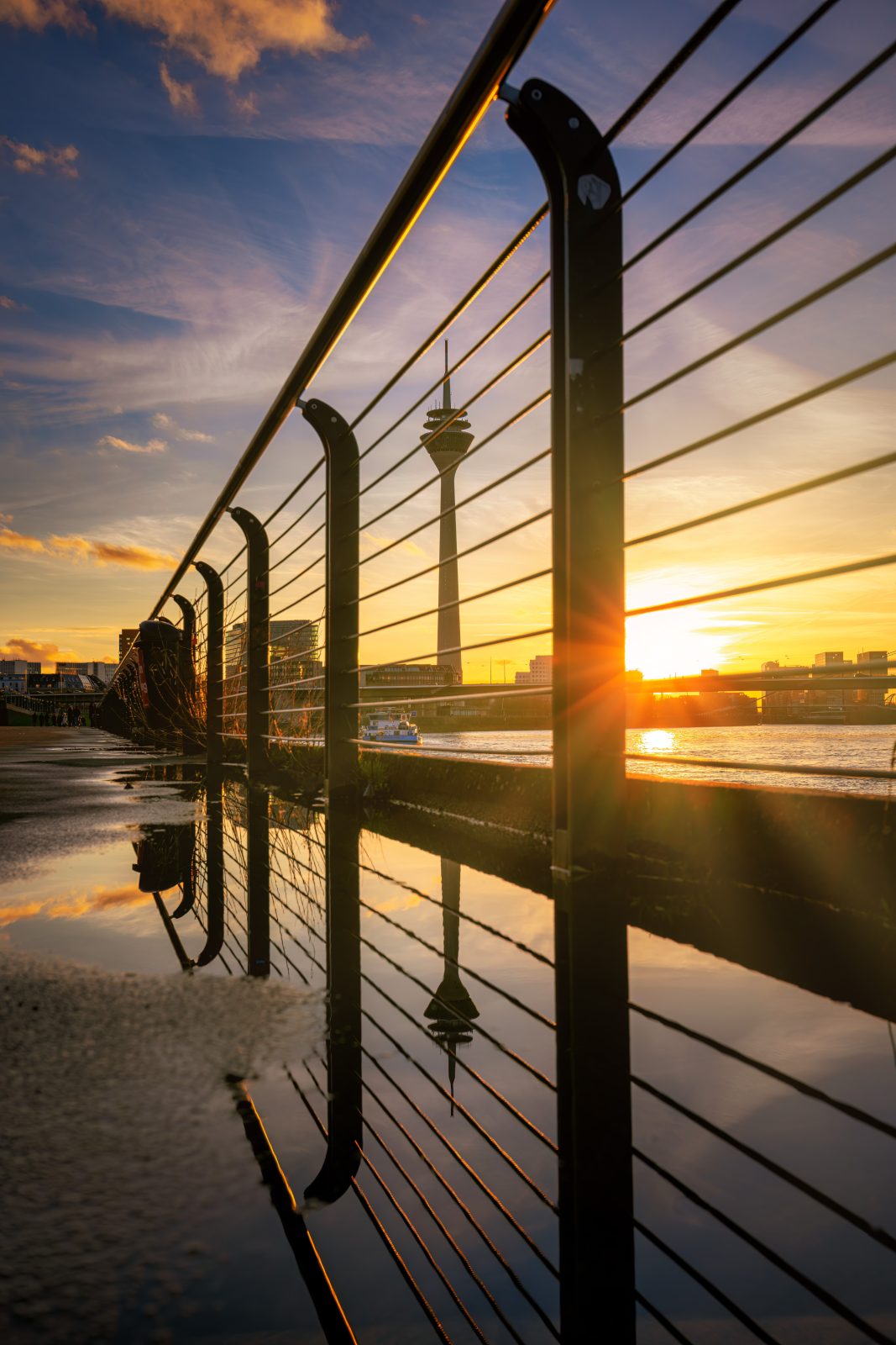 Ein Geländer entlang eines Flussufers spiegelt sich in einer Pfütze. Dahinter stehen eine Brücke, ein Wasserturm und städtische Gebäude im warmen Licht der tief stehenden Sonne.