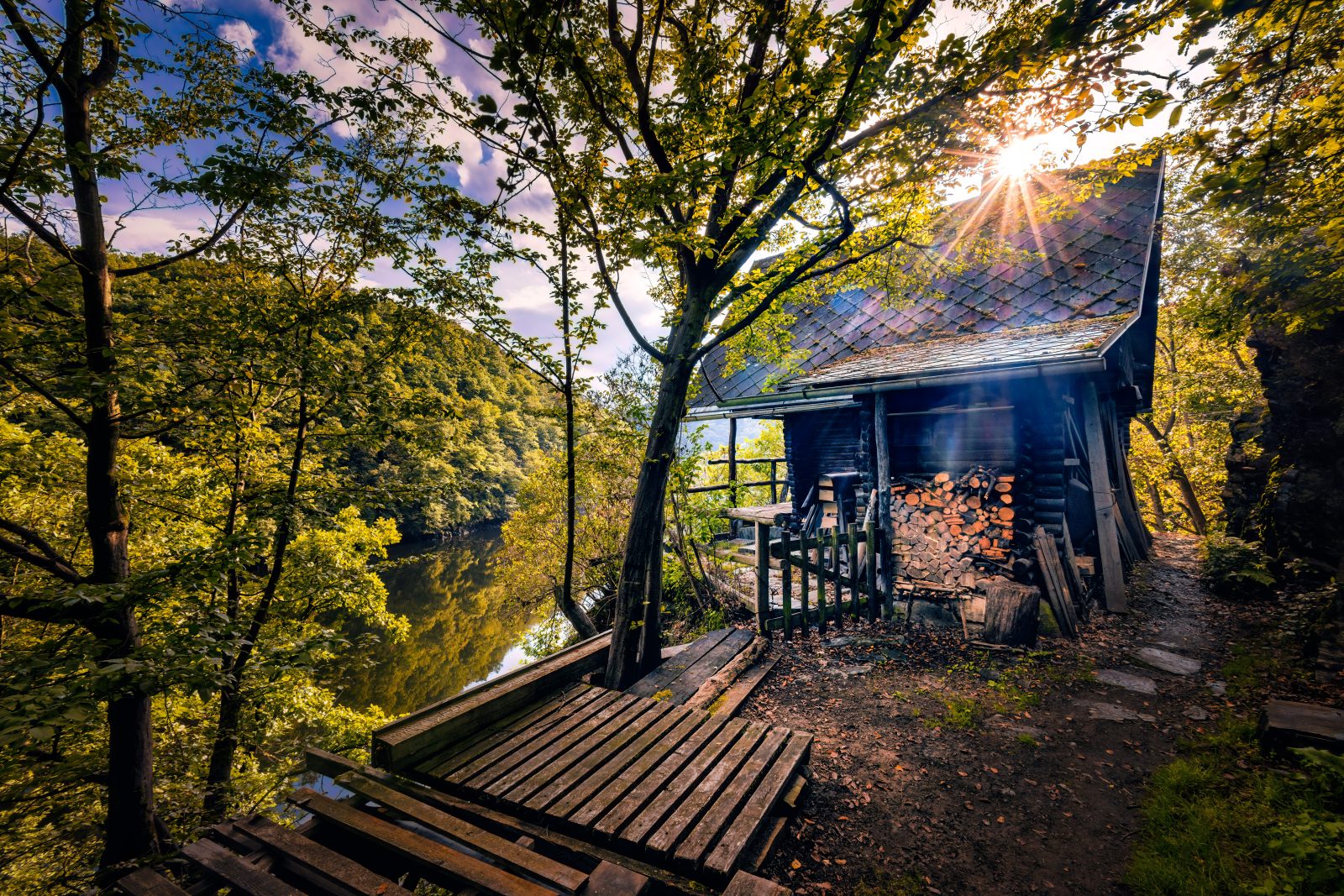 Eine hölzerne Hütte mit gestapeltem Brennholz steht an einem bewaldeten Teich. Eine Holzplattform führt zum Wasser, durch das Blätterdach fällt tief stehendes Sonnenlicht.