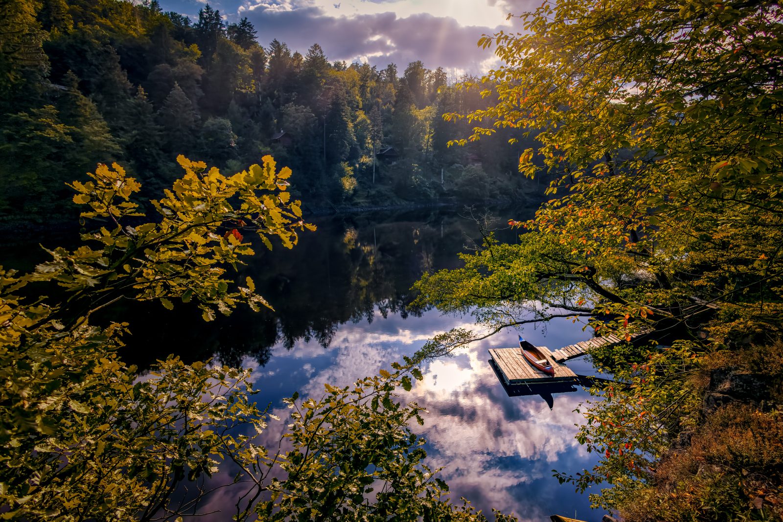 Ein stiller Waldsee mit einem kleinen Steg und Ruderboot, umgeben von herbstlich gefärbten Bäumen; Wolken spiegeln sich auf der Wasseroberfläche.